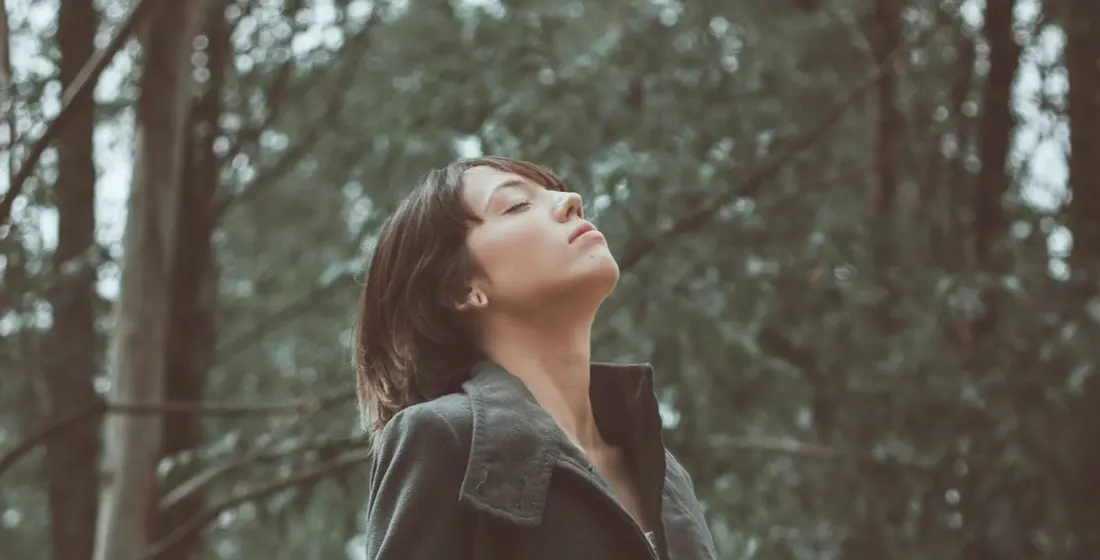 Woman standing among tall trees with her eyes closed, taking a slow deep breath of fresh air